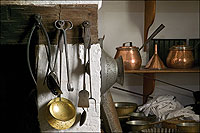 Utensils made of copper, brass, tin, and iron hang from the lintel of a kitchen fireplace.