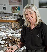 Curator and author Bly Straube stands holding the pendant in front of a table full of objects found in the same location.