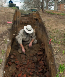 An archaeologist crouches down to examine brick rubble.